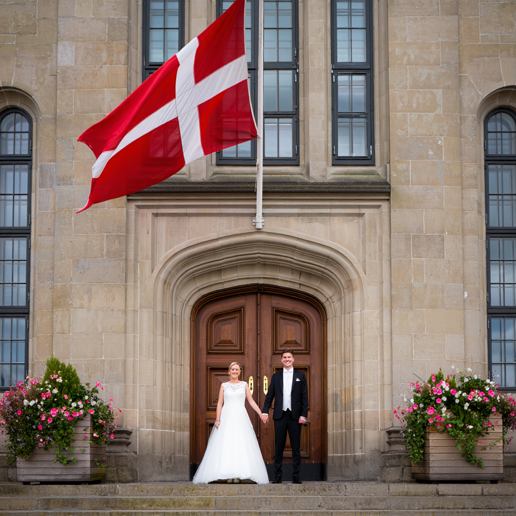 International couple getting married in front of a Danish town hall with Danish flag, symbolizing easy and romantic weddings in Denmark